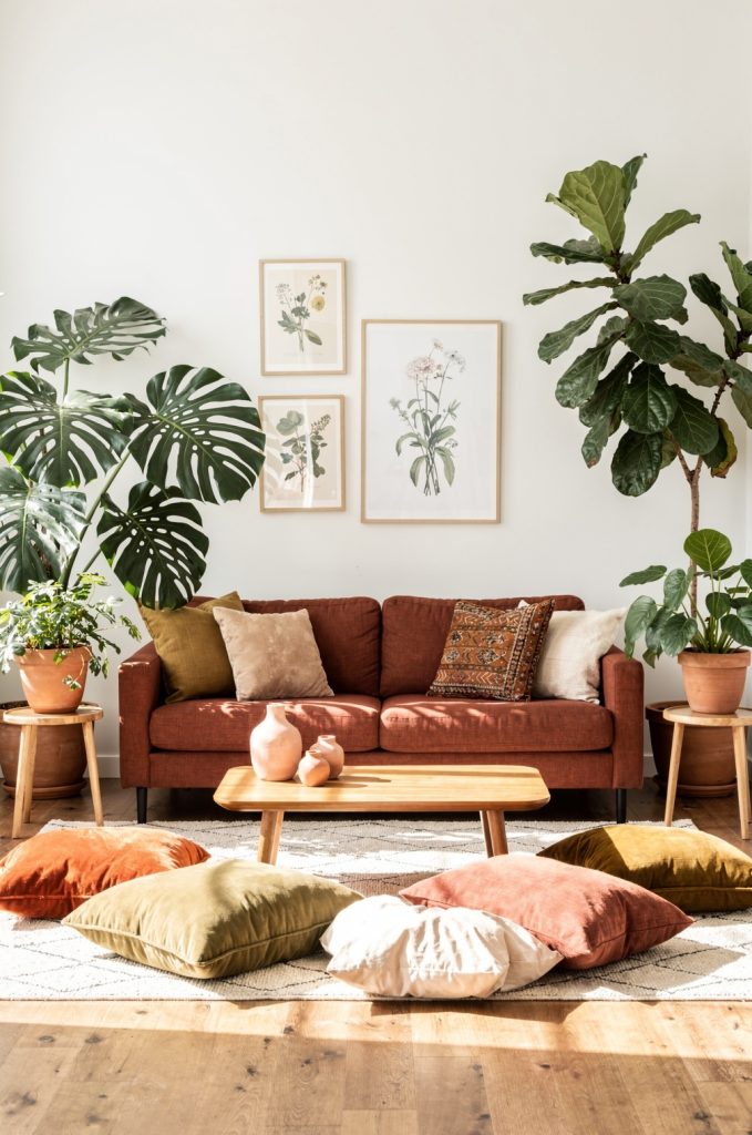Indoor plants including monstera and fiddle leaf fig surrounding a rust sofa in a boho living room with floor cushions and wood furniture.