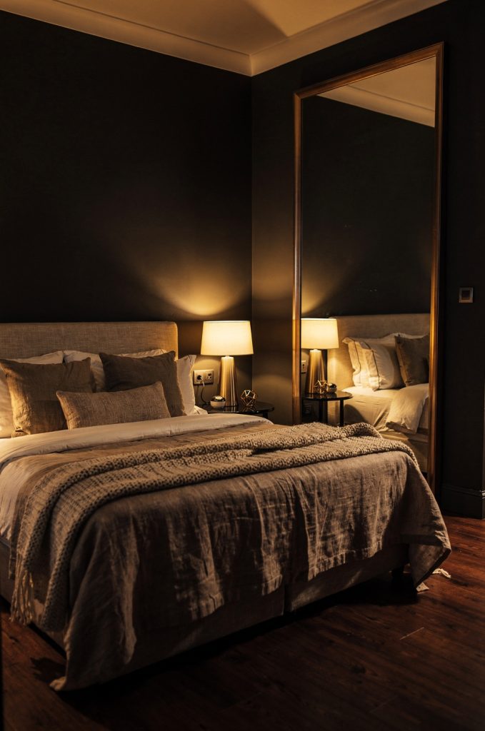 Bedroom with full-length mirror reflecting bed and warm lighting, creating depth and layered visual interest.