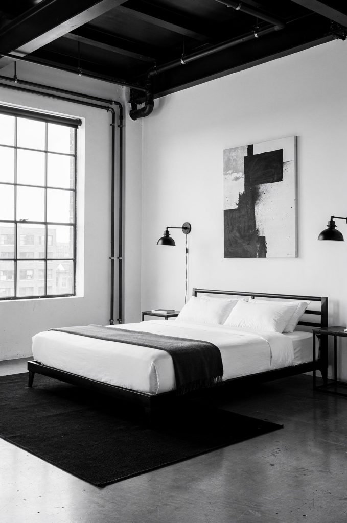 Black and white industrial bedroom with metal bed frame, exposed ceiling beams, wall lamps, and minimal decor.