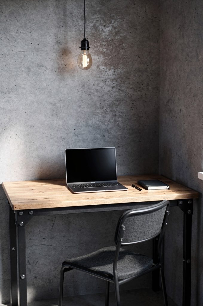 Small industrial workspace with wooden desk, metal frame, pendant light, and concrete wall background.
