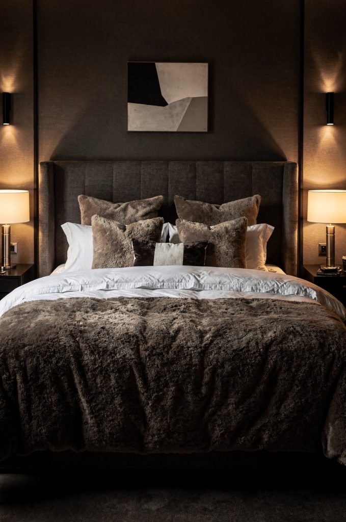 Moody bedroom with winged upholstered headboard, layered pillows, and warm bedside lighting creating a cozy boutique-style atmosphere.