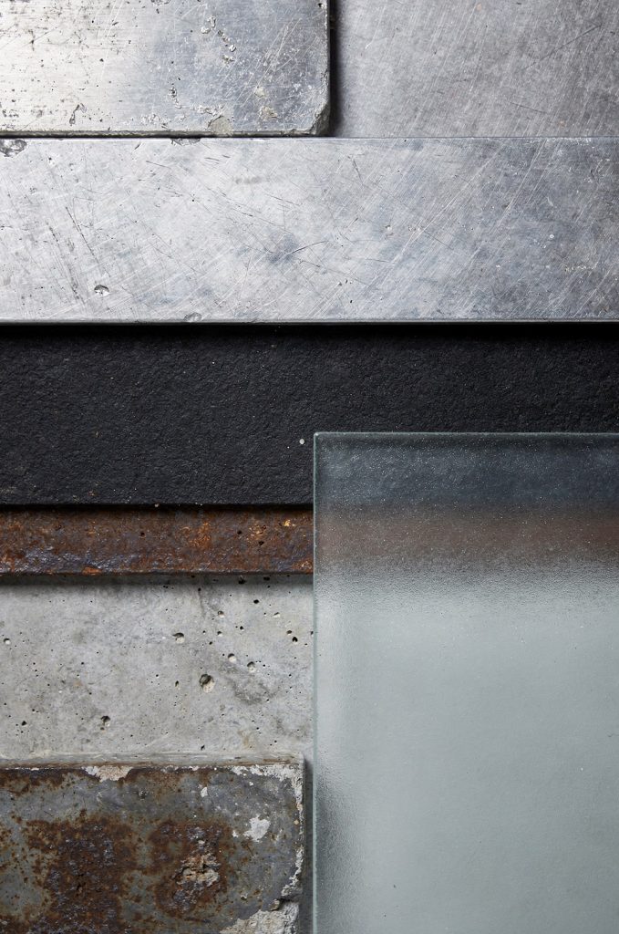 Close-up of industrial materials including brushed metal, rusted steel, concrete, and frosted glass arranged in a layered composition.